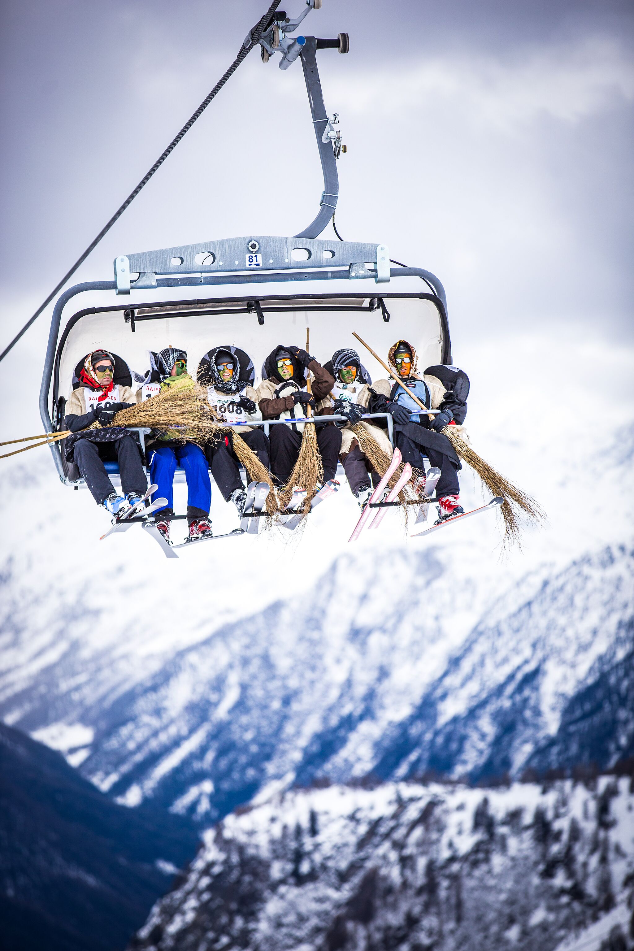 A ski lift with passengers holding brooms, set against a snowy mountain backdrop.