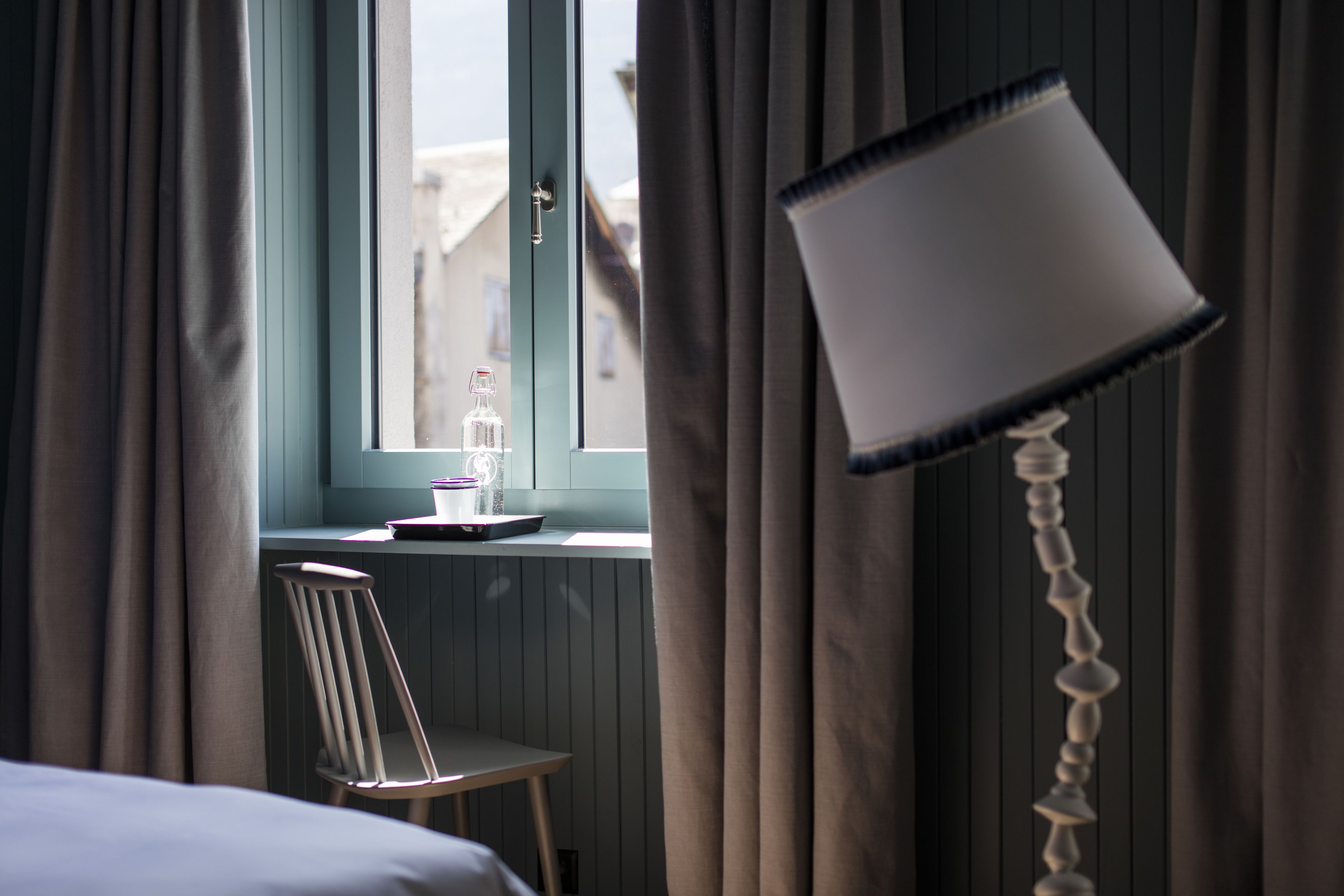 Une pièce avec un lit, une chaise, une fenêtre avec des rideaux et une lampe.