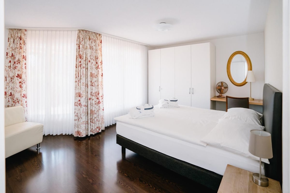 Quarto de hotel com cama, mesa de cabeceira, cadeira, guarda-roupa e espelho.