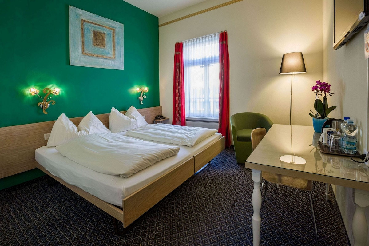 A hotel room with a double bed, a desk with a lamp, a chair, a window with red curtains, and wall-mounted lights.