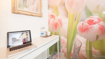 Una computadora portátil sobre un escritorio, con un espejo encima, un florero de vidrio con una tulipa, y un fondo borroso de flores.