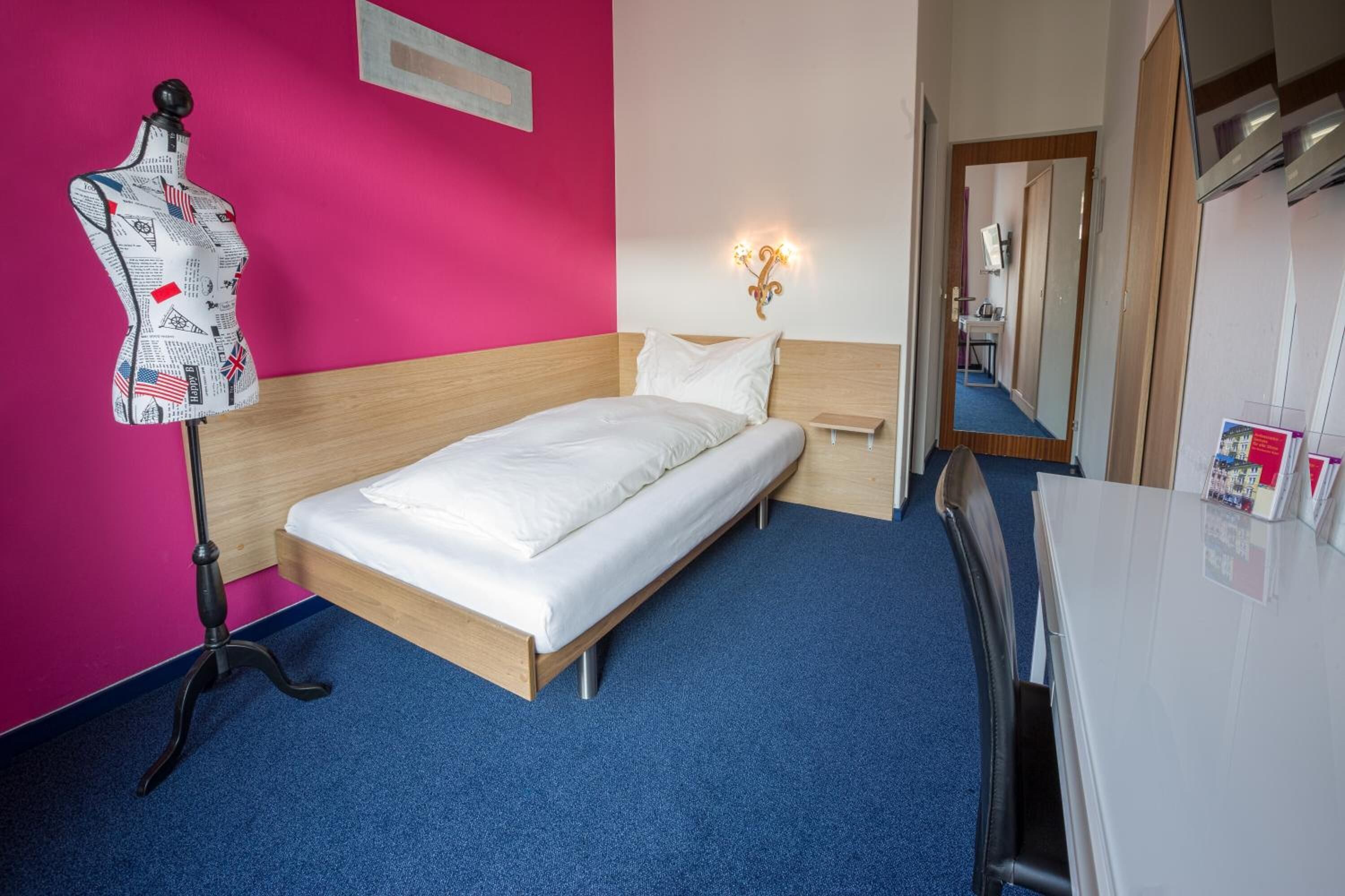 A hotel room with a bed, a mannequin, a nightstand, a mirror, and a desk.