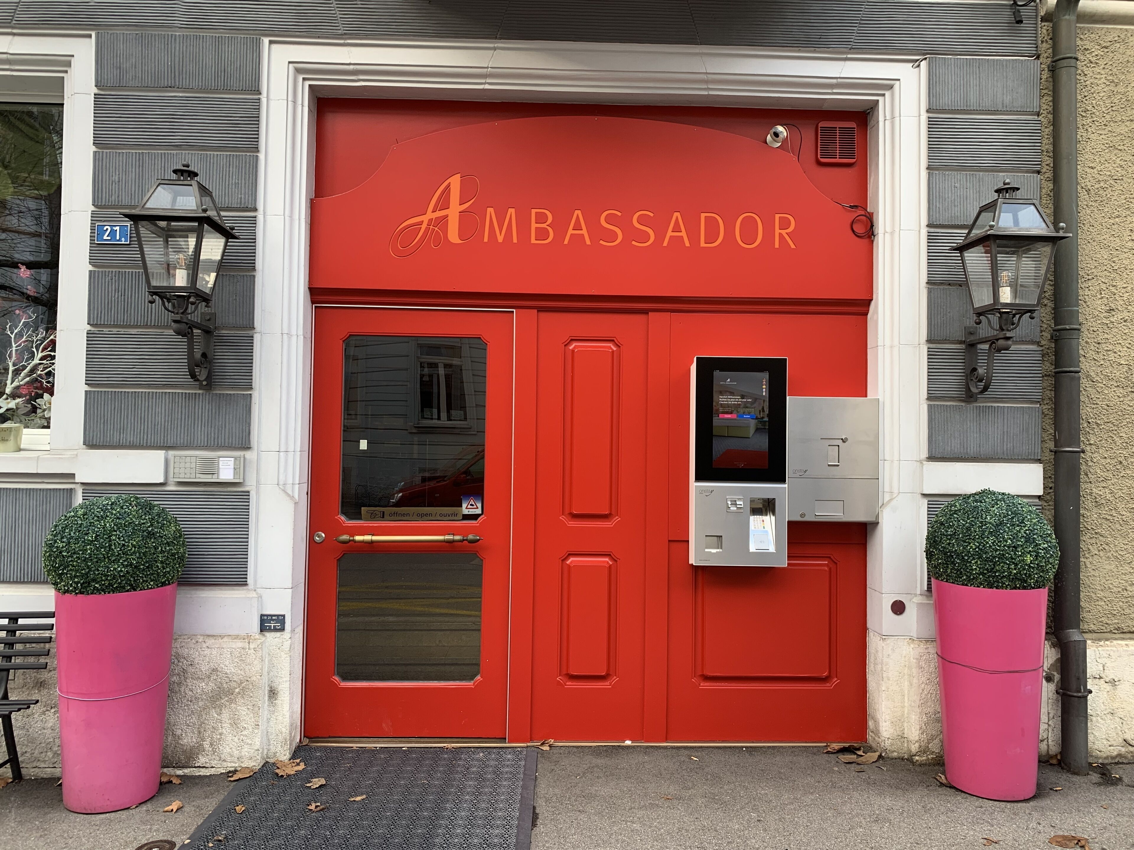 A red double door with the word "AMBASSADOR" above it, a keypad entry system, and two potted plants on either side.