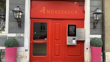 A red double door with the word "AMBASSADOR" above it, a keypad entry system, and two potted plants on either side.