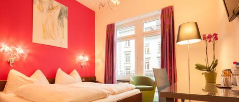 A hotel room with a bed, a desk, a chair, a potted plant, and a window with curtains.