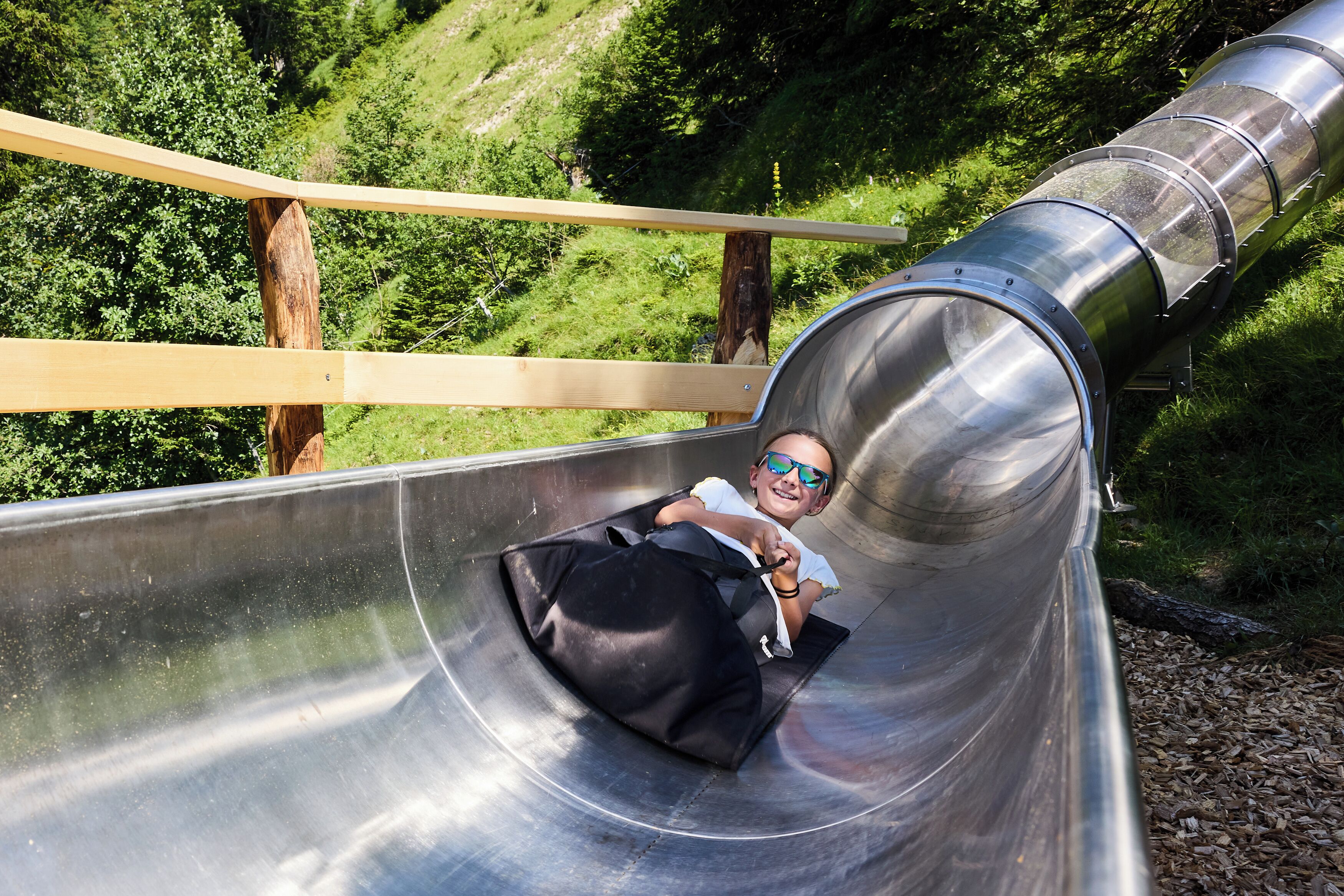 A person sliding down a metal slide with a black bag, surrounded by greenery.
