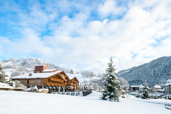 A wooden lodge with a large glass facade, surrounded by snow-covered mountains and trees. - Hotel Cailler (Charmey)