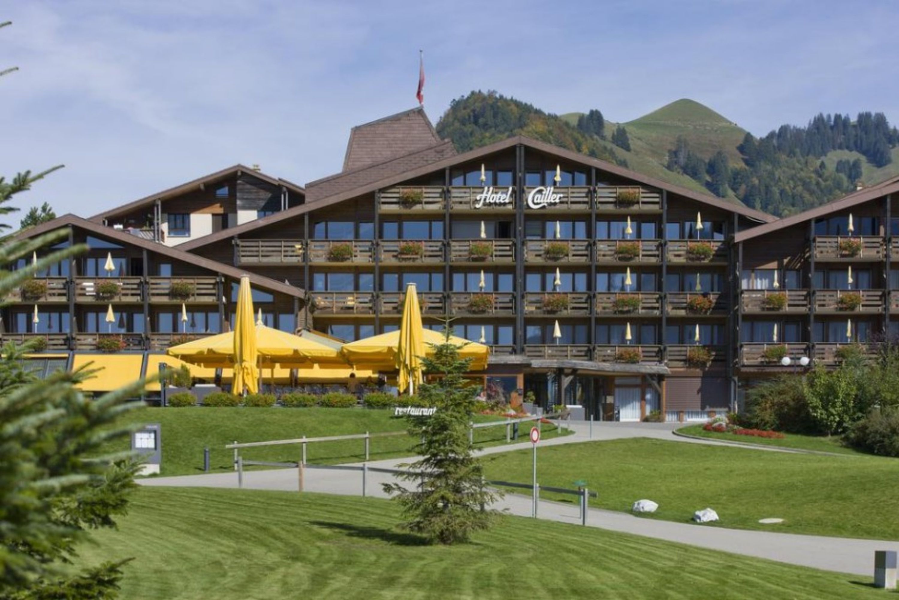 Ein mehrstöckiges Hotel mit Balkonen, einem zentralen Eingang und einer umlaufenden Rasenfläche.
