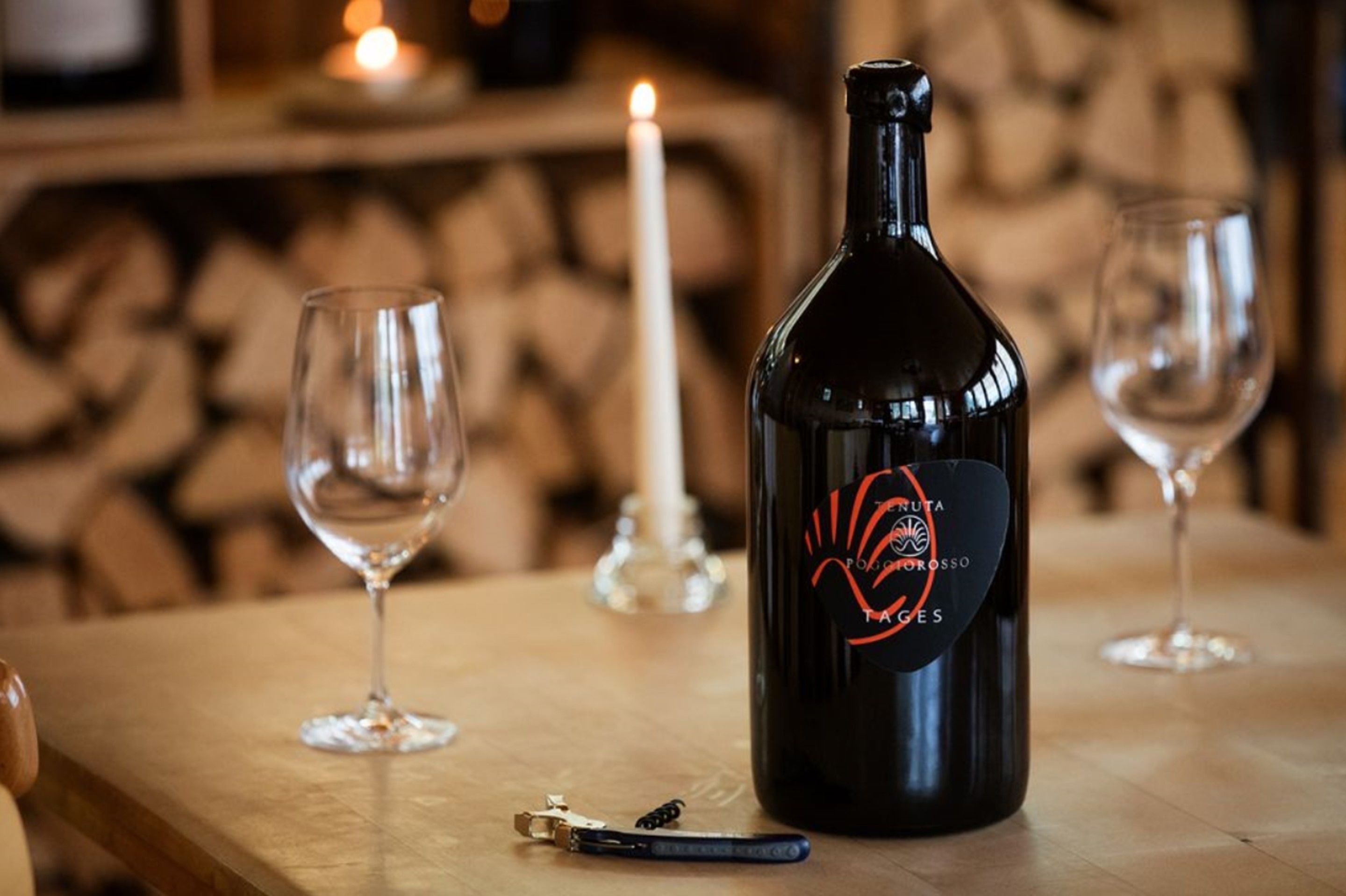 Una botella de vino con un sacacorchos sobre una mesa de madera, dos copas de vino vacías y una vela encendida al fondo.