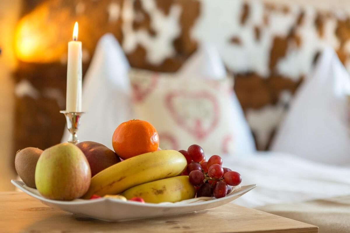 Un plato de fruta con manzanas, plátanos, uvas y una naranja, con una vela encendida al fondo.