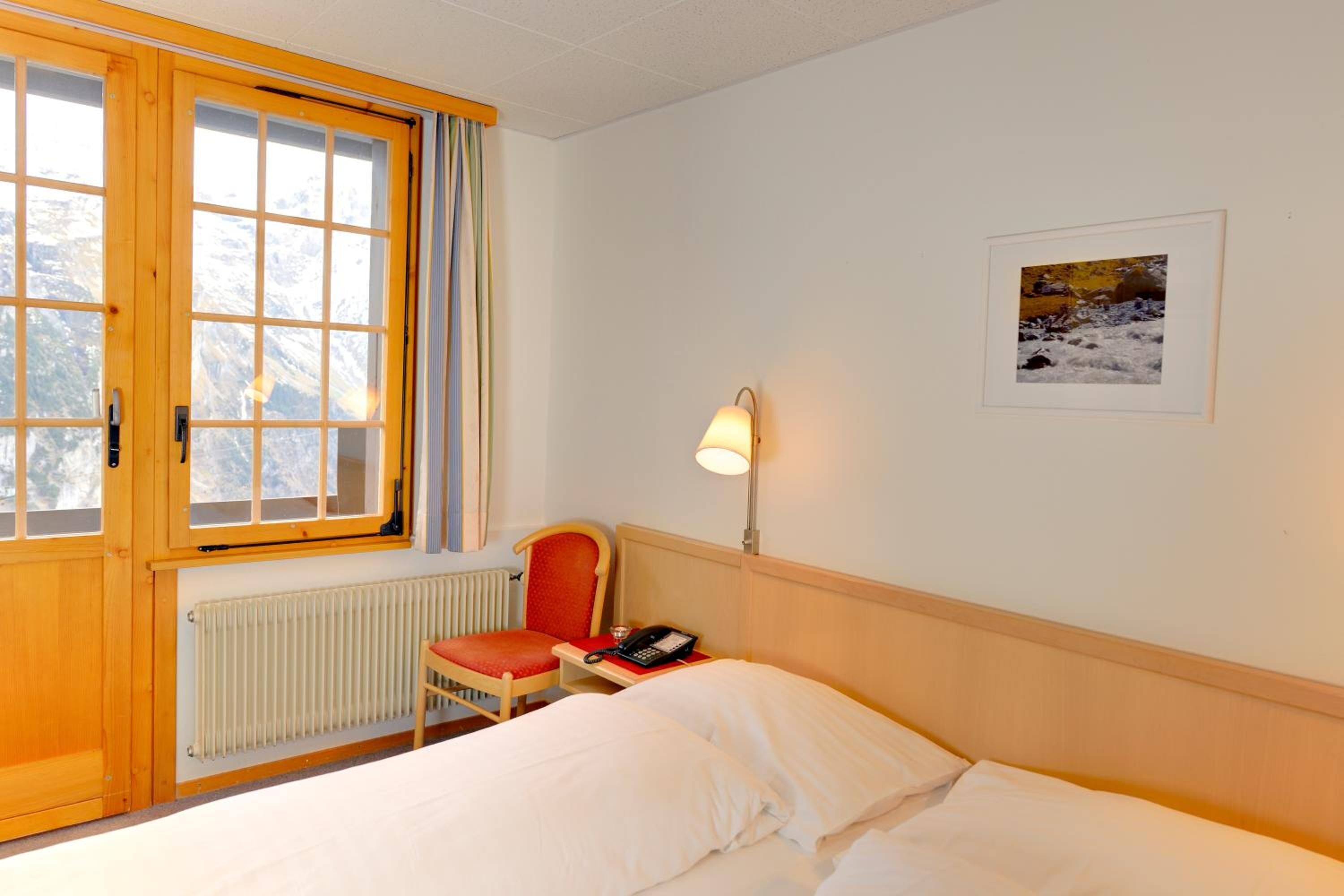 Habitación de hotel con cama, una silla, escritorio con teléfono, lámpara, cuadro en la pared y ventana con cortinas.