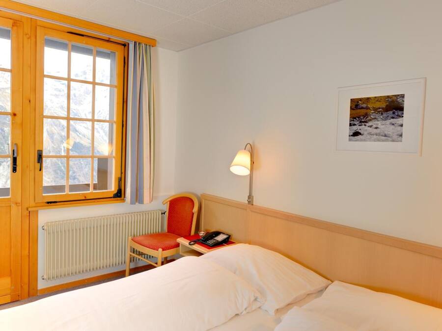 Habitación de hotel con cama, una silla, escritorio con teléfono, lámpara, cuadro en la pared y ventana con cortinas.