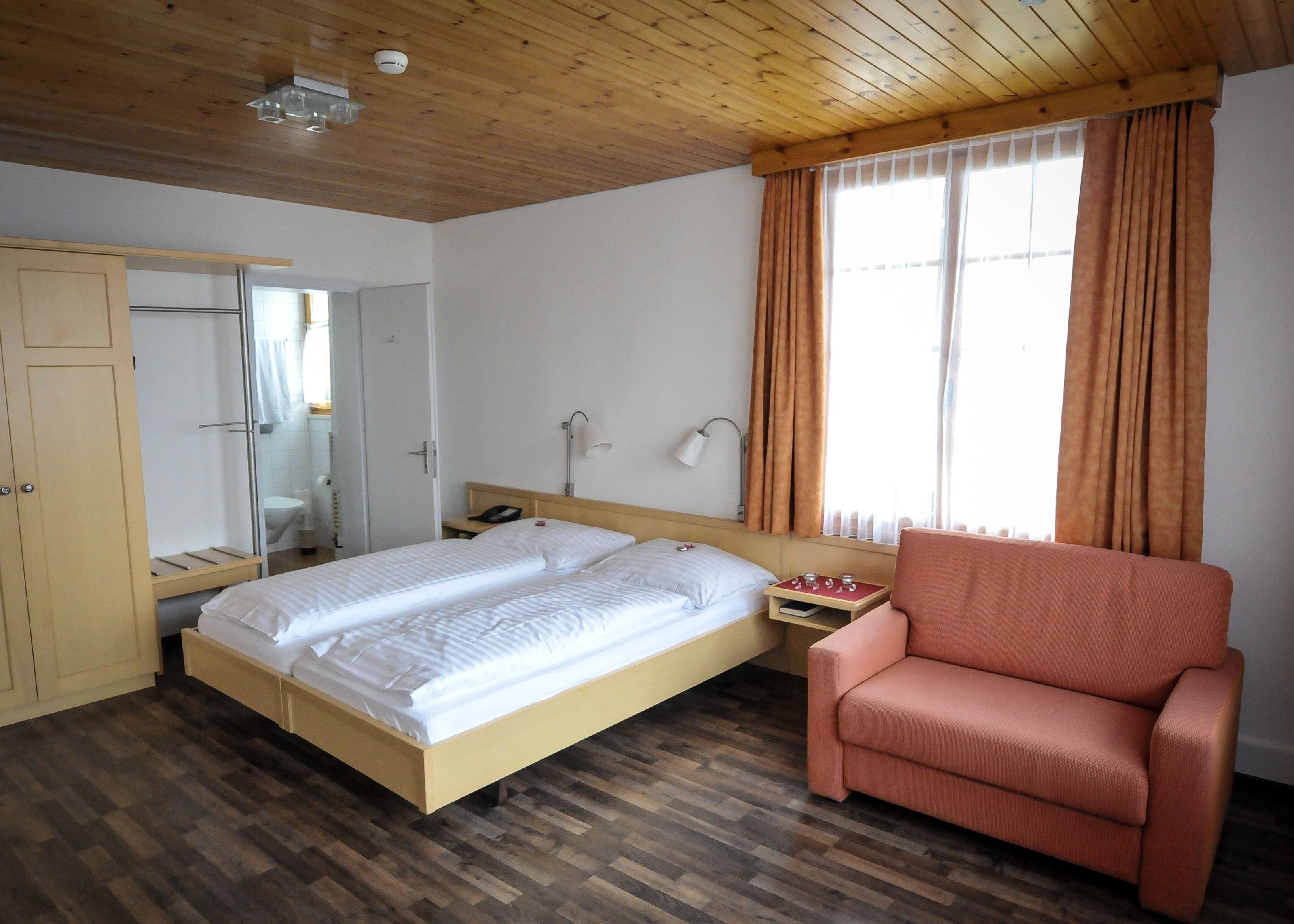 A hotel room with a wooden ceiling, a bed with white linens, a wooden nightstand, a red chair, and a window with curtains.