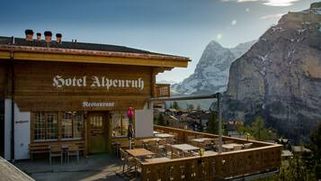A wooden building with a sign reading "Hotel Alpenruh Restaurant." It features outdoor seating with tables and chairs, and a view of snow-capped mountains in the background.