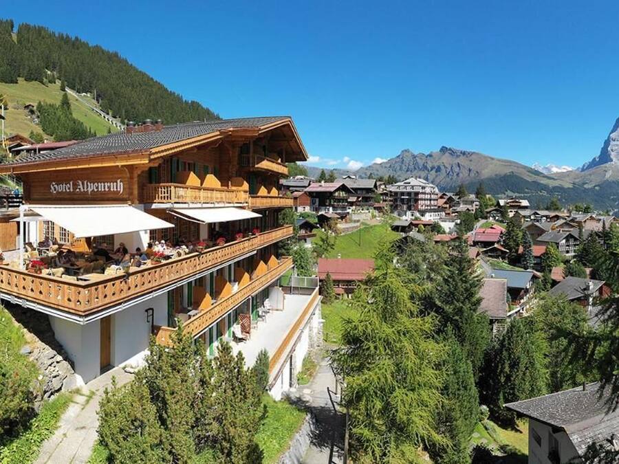 Un hotel con fachada de madera, mobiliario exterior y vistas a un pueblo de montaña.