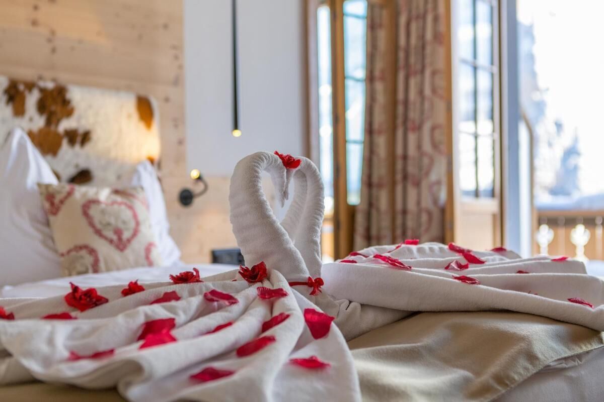A bed with a white blanket and red rose petals, a heart-patterned pillow, and a rustic wooden headboard.