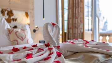 A bed with a white blanket and red rose petals, a heart-patterned pillow, and a rustic wooden headboard.