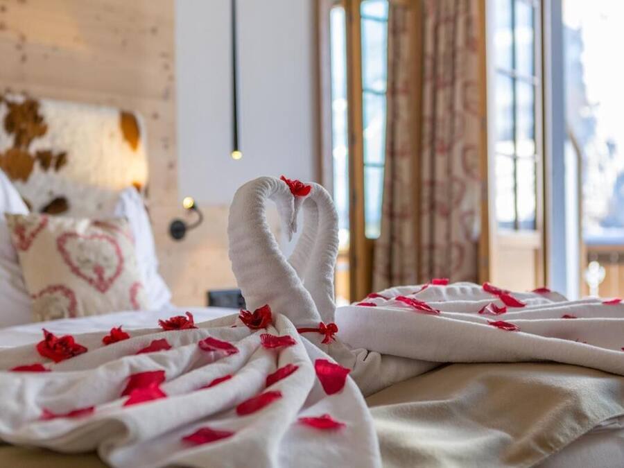 Una cama con una colcha blanca y pétalos de rosa roja, un cojín con forma de corazón y un cabecero de madera rústica.