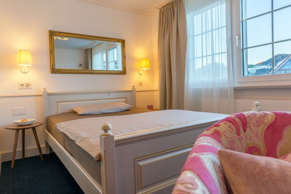 A neatly made bed with a white headboard, a bedside table with a bowl of fruit, and a window with curtains. - Hotel Appenzell (Appenzell)