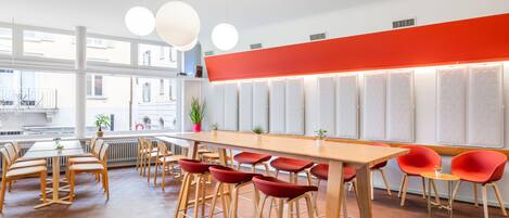 A modern restaurant with a long wooden table and red chairs, large windows, and a red accent wall.