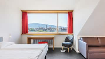 Une chambre d’hôtel avec un lit, un bureau, une chaise et un canapé. Il y a une grande fenêtre avec des rideaux rouges, d’où l’on peut admirer des bâtiments et des collines.