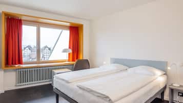 Ein Hotelzimmer mit Bett, Sessel, Schreibtisch, Lampe und einem fensterbelagten Fenster.