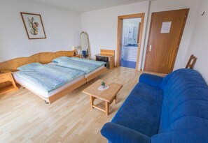 A bedroom with a bed, a blue sofa, a wooden coffee table, a dresser, a mirror, and a door leading to a bathroom. - Hôtel Restaurant Le Gruyerien (Morlon)