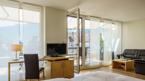 A modern hotel room with a large bed, a desk with a TV, a chair, a sofa, and a balcony with a view of buildings and mountains. - Hotel Artos Interlaken (Interlaken)