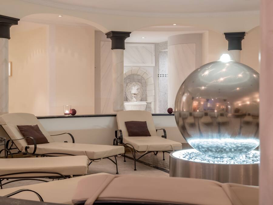 Ein Wellnessbereich mit Liegestühlen, einer Zierbrunnen und einer Marmorwand mit einer Löwenstatue.