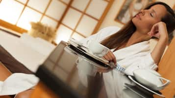 A person in a white robe holding a tray with a cup and saucer, a bowl, and a spoon, in a room with a wooden floor and a large window with a grid pattern.