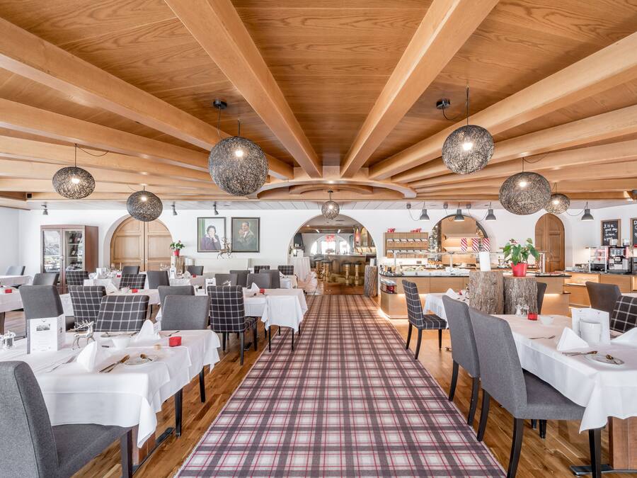 Um restaurante com vigas de madeira no teto, luminárias pendentes redondas e um tapete xadrez que conduz a mesas com toalhas brancas e cadeiras.