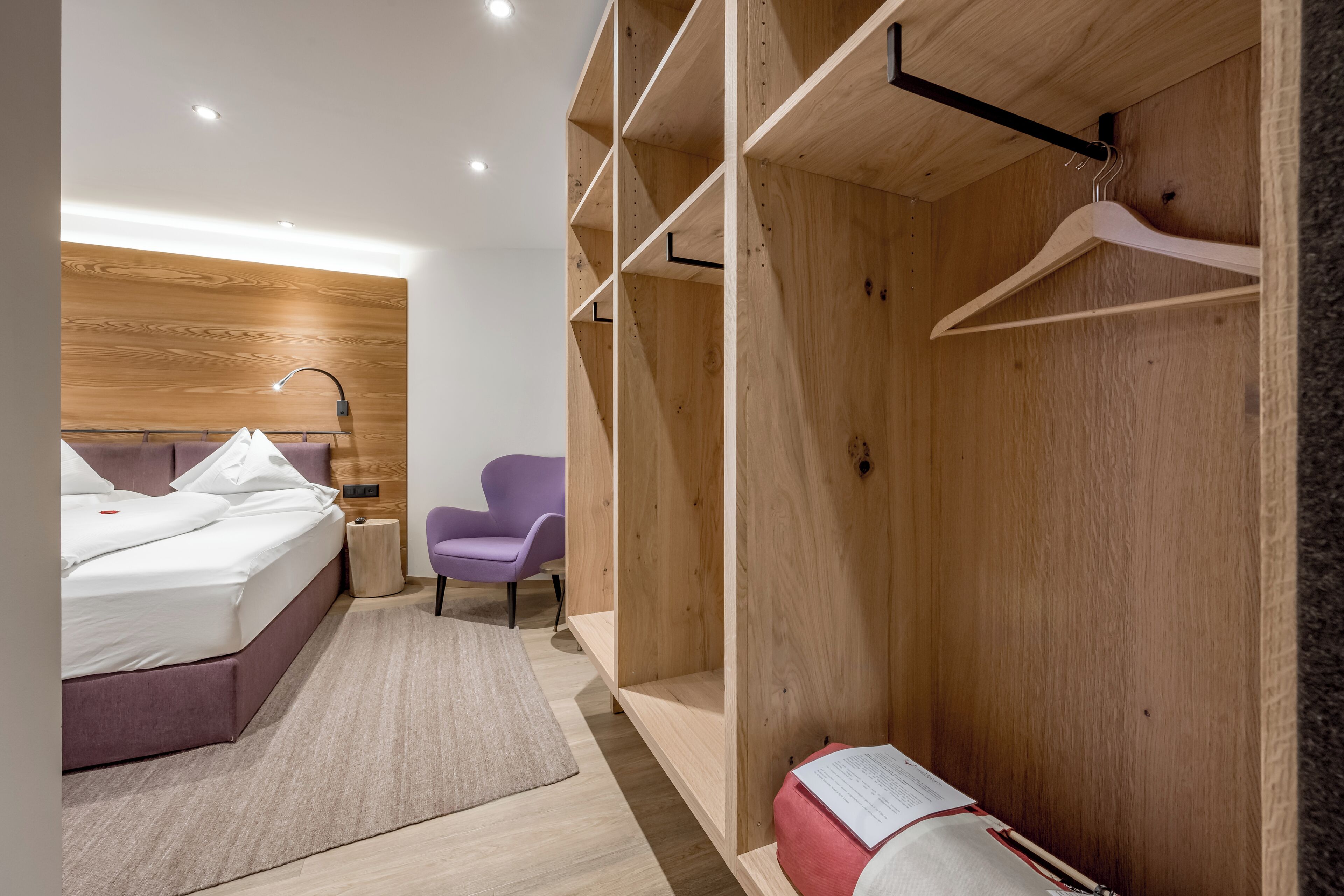 Une chambre d’hôtel moderne avec une tête de lit en bois, un lit, un fauteuil violet, une armoire en bois et une petite table sur laquelle se trouve un sac rouge.