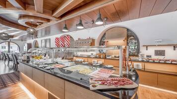 Zona de buffet con diversas opciones de comida, área de comedor con mesas y sillas, y diseño interior moderno con vigas de madera e iluminación colgante.