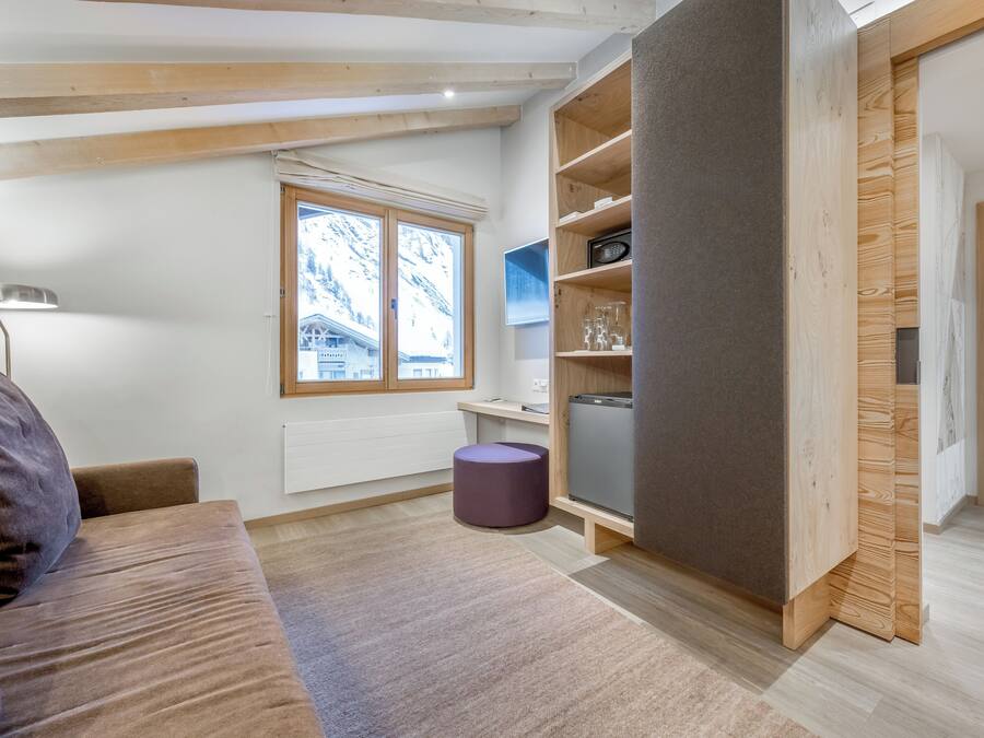 Ein moderner, hell eingerichteter Raum mit großem Fenster, Holzbalken, einer Couch, einem kleinen Tisch und einem violetten Pouf.