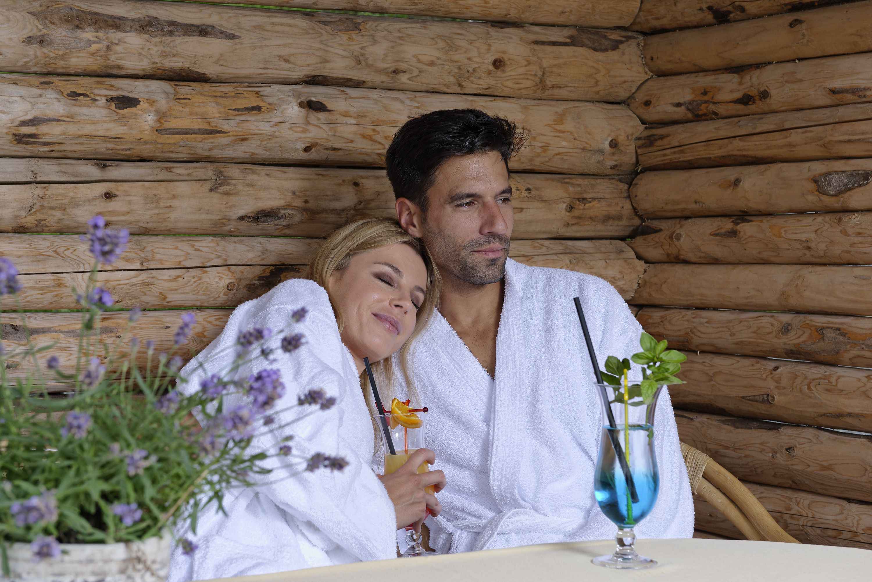 A couple in white robes seated at a wooden table with a drink and flowers.