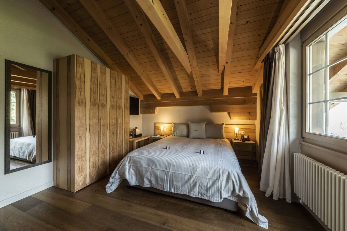 Une chambre à coucher avec un grand lit, des poutres apparentes en bois au plafond, une table de chevet avec une lampe et une fenêtre avec des rideaux.