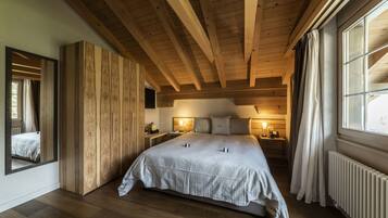 A bedroom with a large bed, wooden ceiling beams, a nightstand with a lamp, and a window with curtains.