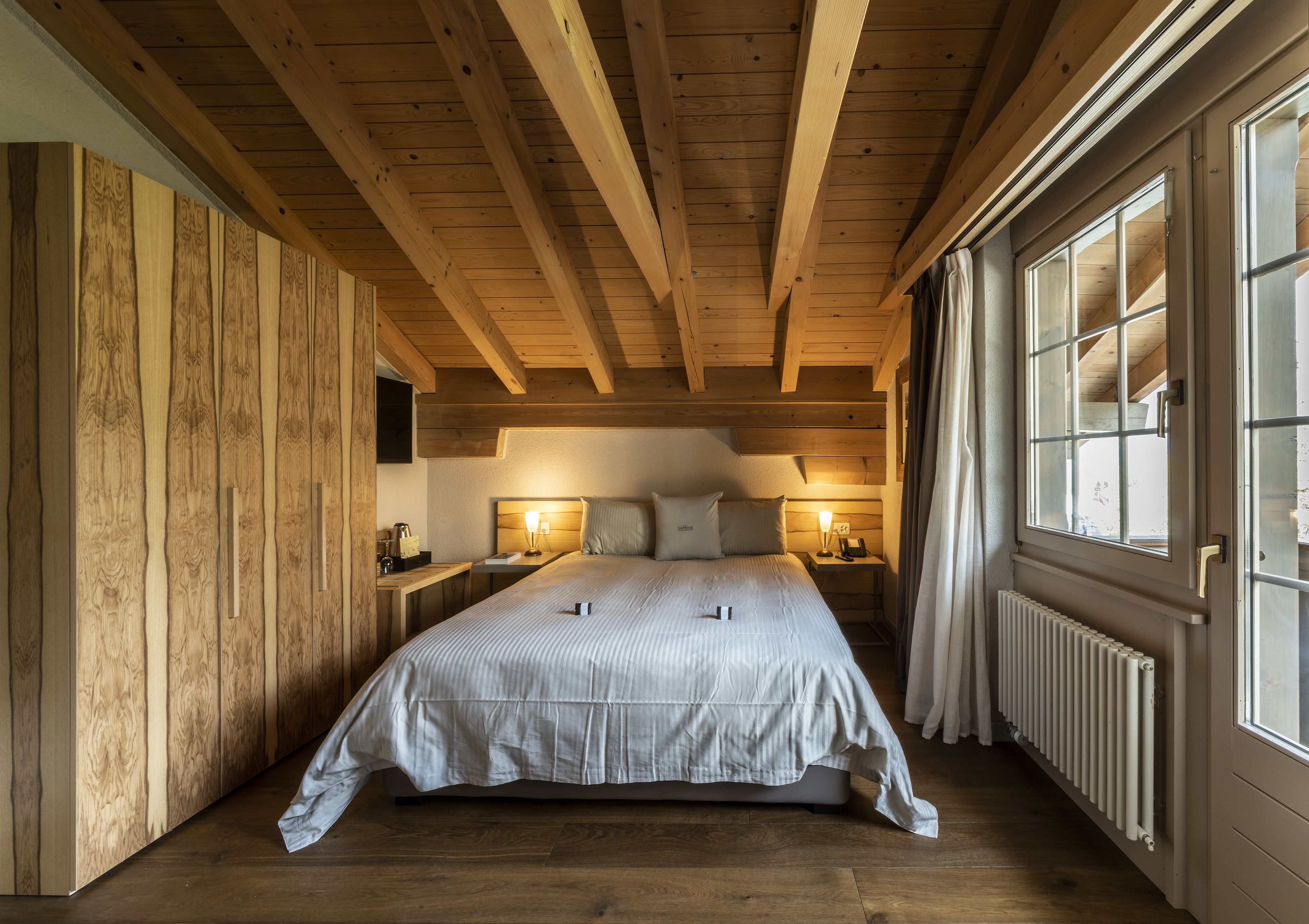 A cozy bedroom with a wooden ceiling, a large bed, bedside lamps, and a window with curtains.