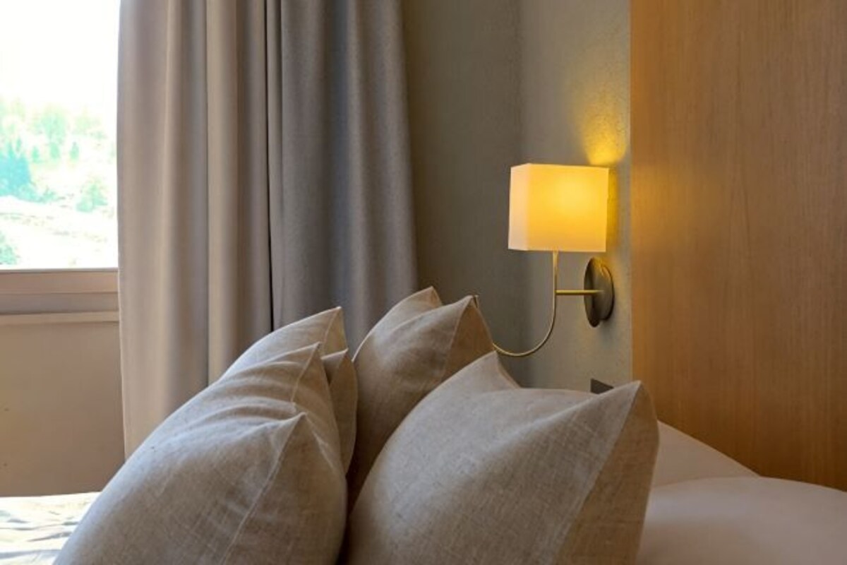 A hotel room with a bed, pillows, a wall-mounted lamp, and a window with curtains.