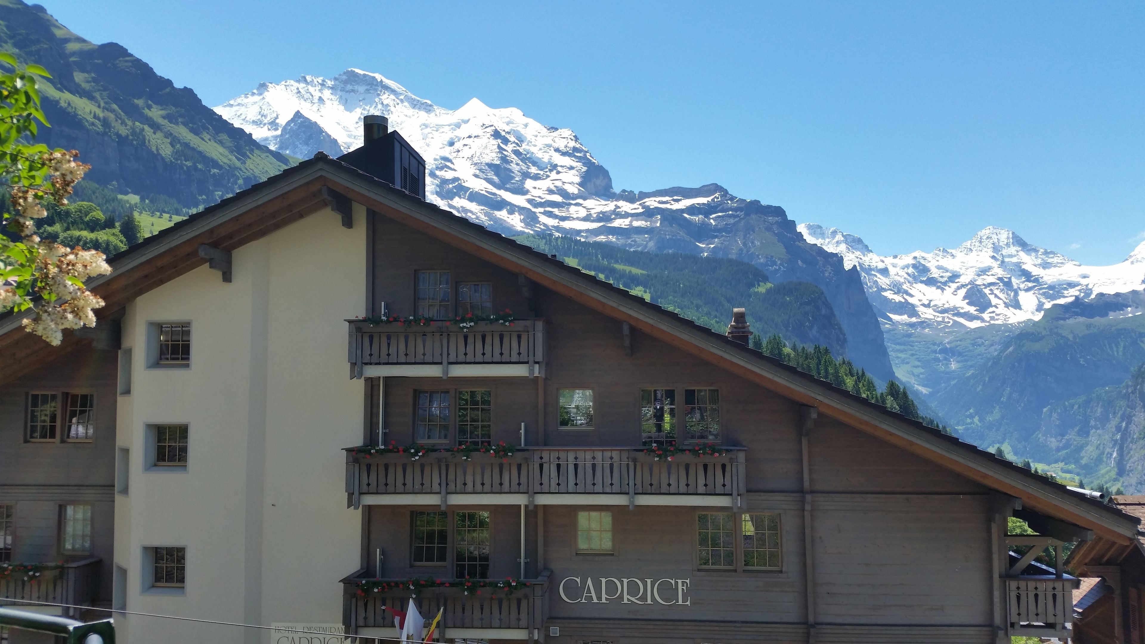 A hotel with a mountain backdrop, featuring balconies with flower arrangements and the name "CAPRICE" on the facade.