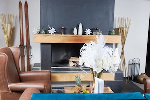 A modern living room with a fireplace, a leather armchair, and a wooden coffee table with fruits and a vase of flowers.