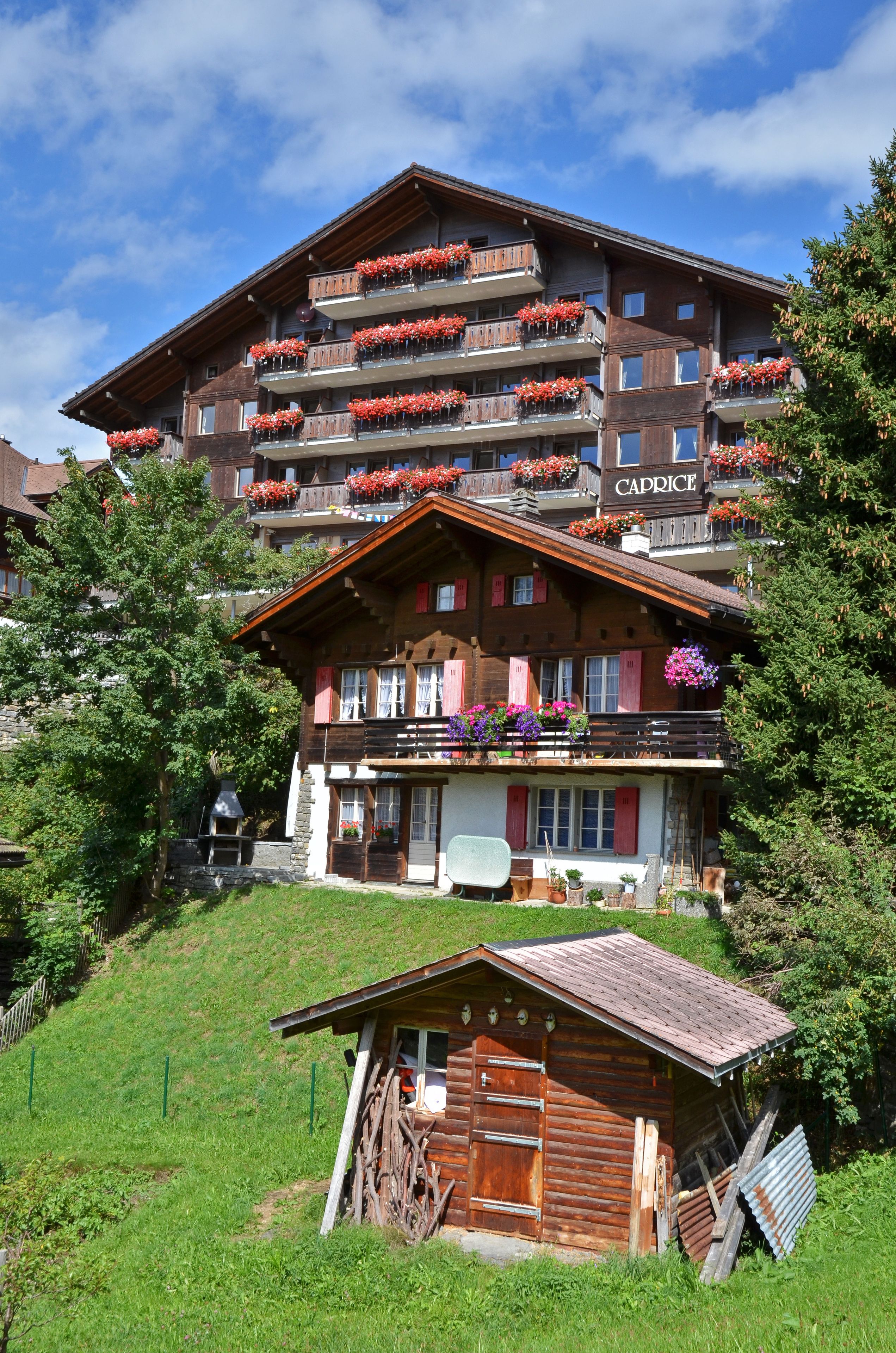 A multi-story building with balconies adorned with flowers, a wooden cabin in the foreground, and a clear blue sky.