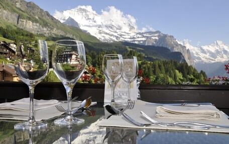 A table set with wine glasses and cutlery, overlooking a mountainous landscape with snow-capped peaks and lush greenery.