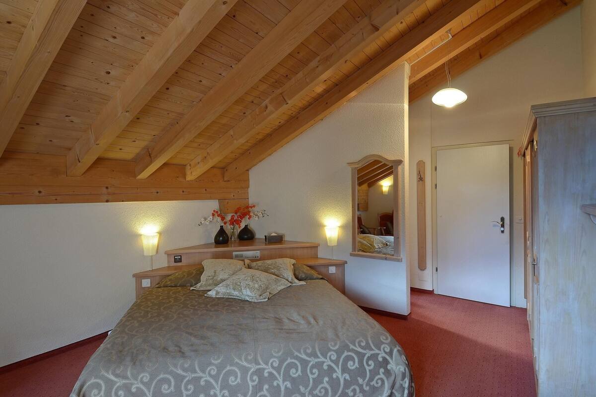 Une chambre avec un plafond en bois, un lit recouvert d’une couvre-lit à motifs, deux lampes fixées au mur, un miroir et une porte.