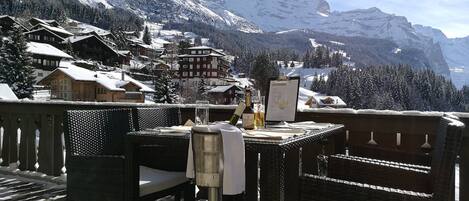 A terrace with wicker furniture, a table with a menu, and a view of snow-covered mountains and buildings.