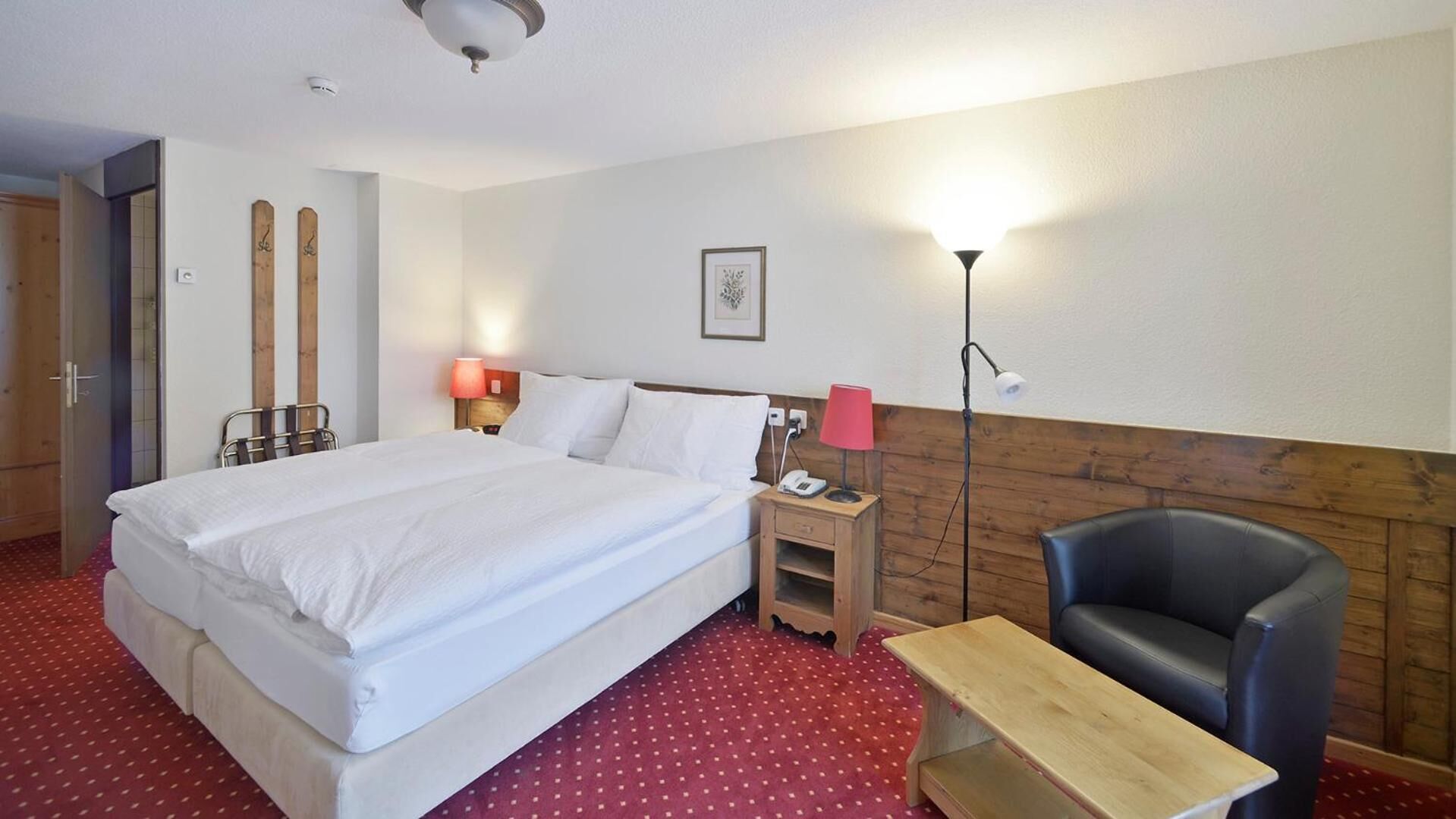 Quarto de hotel com uma cama grande, uma mesa de cabeceira de madeira com uma luminária, uma escrivaninha, uma cadeira e um suporte para esquis.