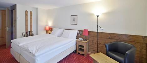 Quarto de hotel com uma cama grande, uma mesa de cabeceira de madeira com uma luminária, uma escrivaninha, uma cadeira e um suporte para esquis.