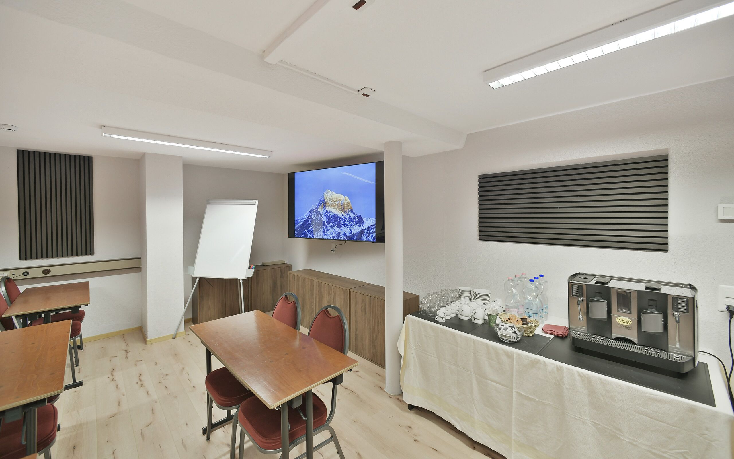 Une salle de conférence équipée d’un tableau blanc, d’un téléviseur diffusant une image de montagne et d’une machine à café.