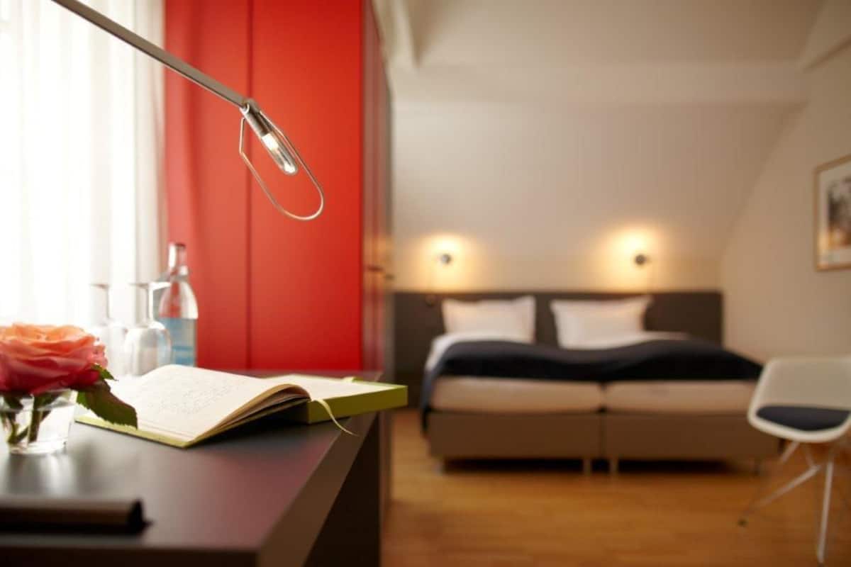 A modern bedroom with a bed, bedside lamps, a desk with a book and a vase of flowers.