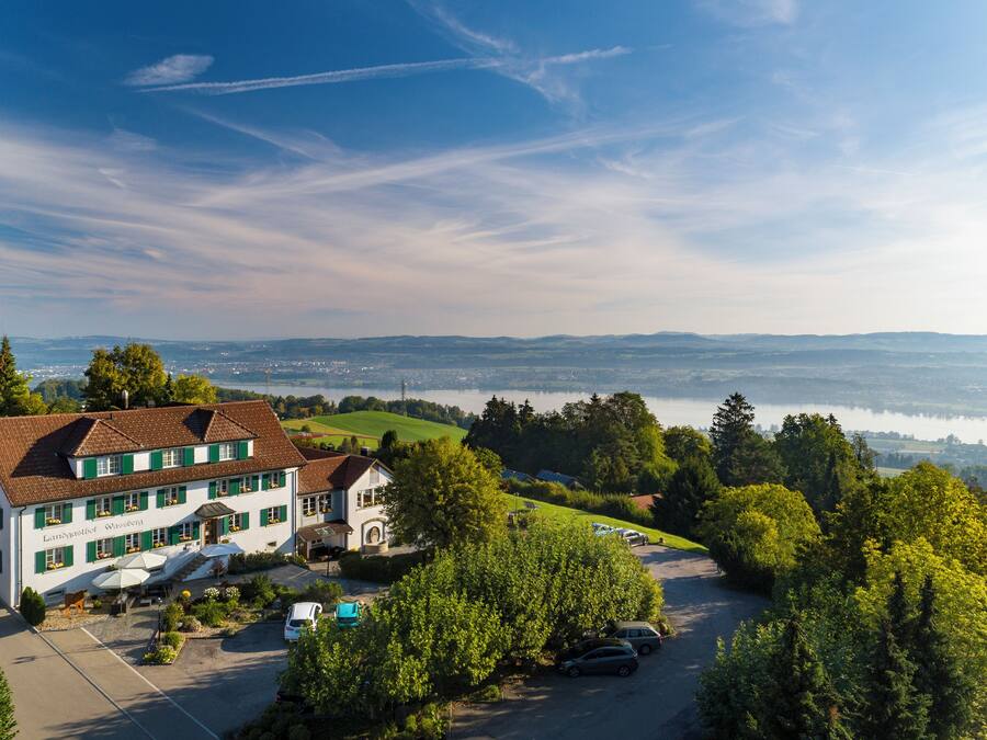 Un edificio blanco grande con contraventanas verdes, un estacionamiento y vistas a un lago y montañas distantes.